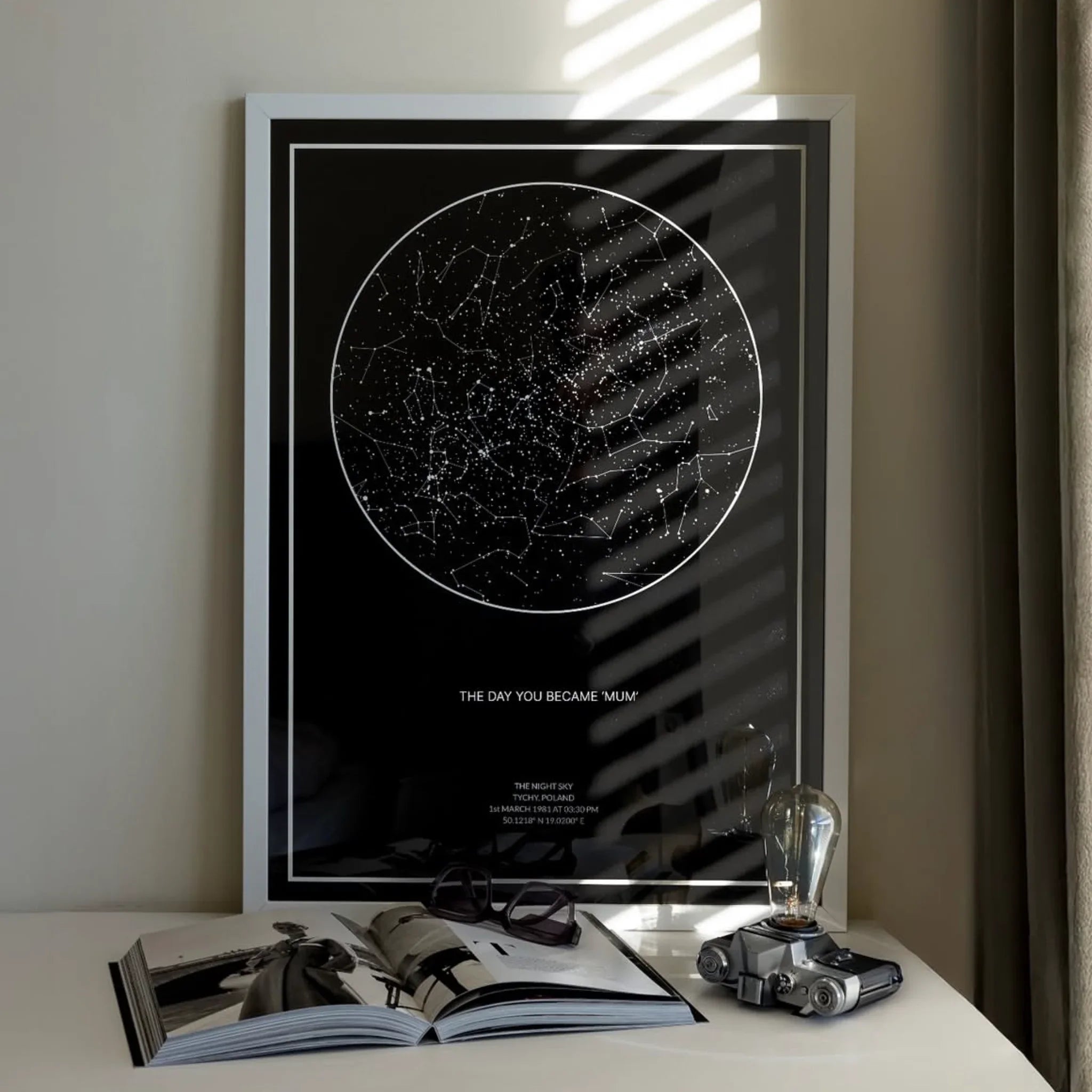 Framed personalized star map print on a tabletop with an open book and decor items
