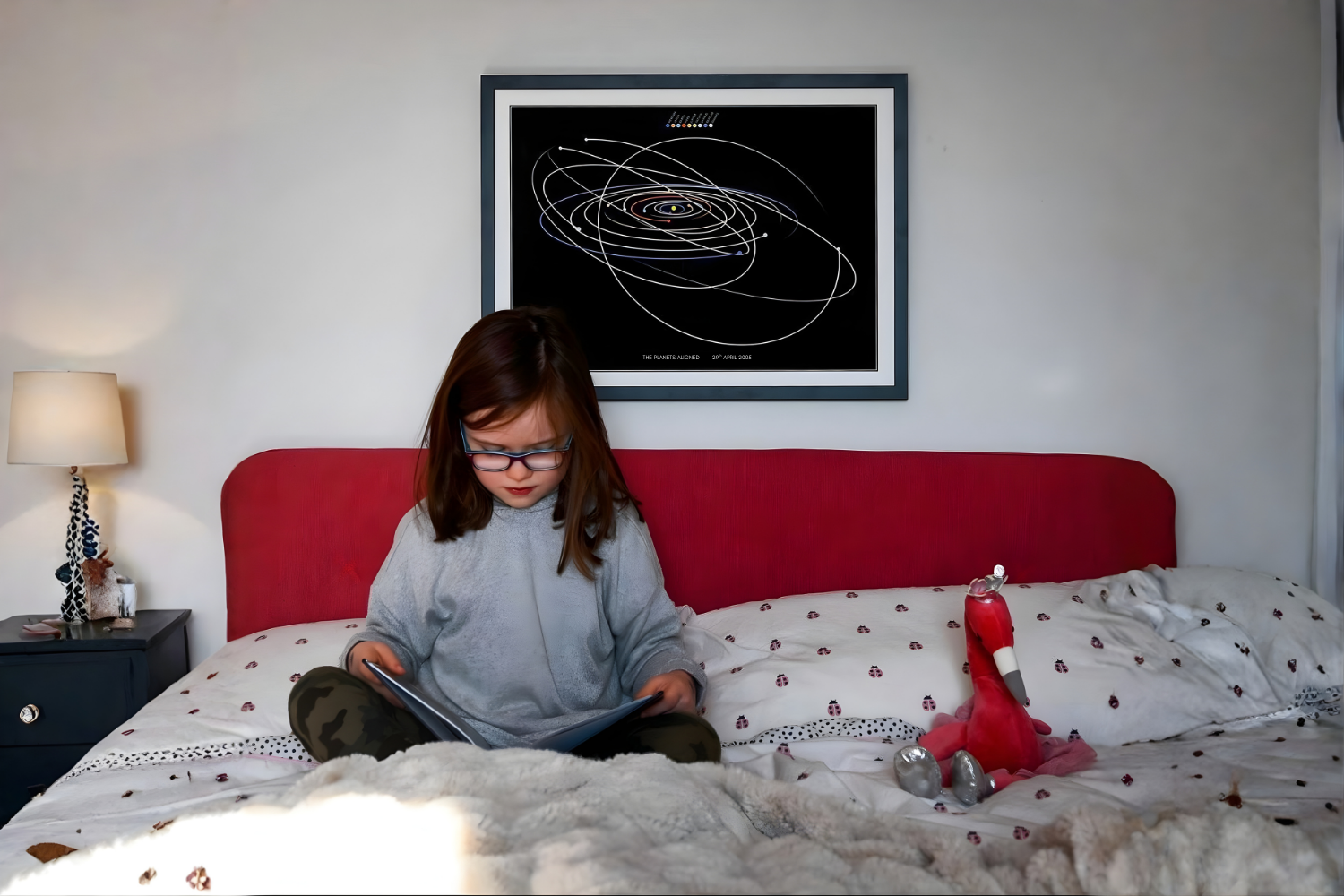 Young girl sitting on bed, reading a book with a framed solar map behind her
